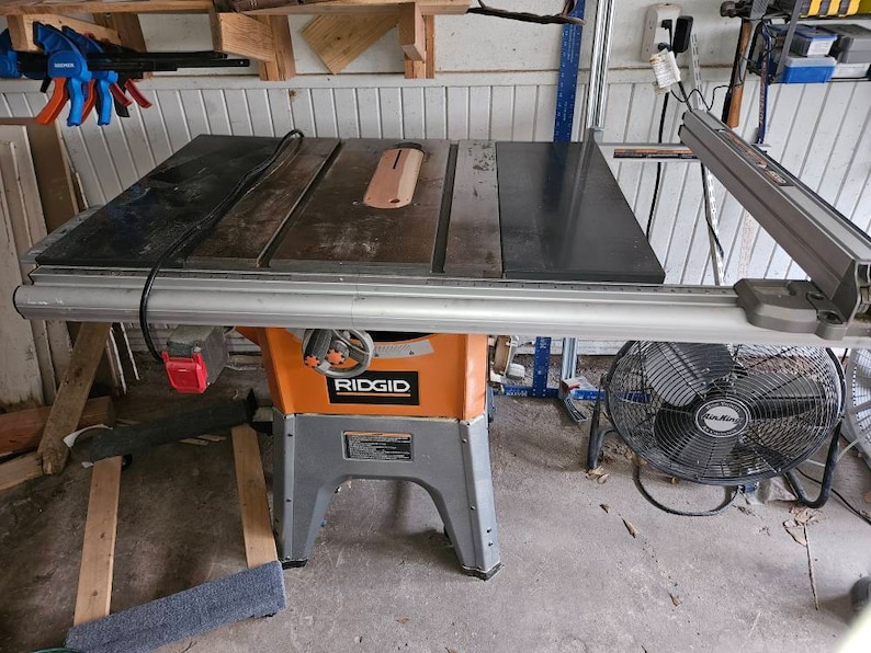 Op de afbeelding: Een Ridgid tafelzaag met een zwart blad en een grijze basis. De zaag heeft een zilveren bladbeschermer en een zwart stroomkabel. De zaag staat op een betonnen vloer. Er is een ventilator op de achtergrond.