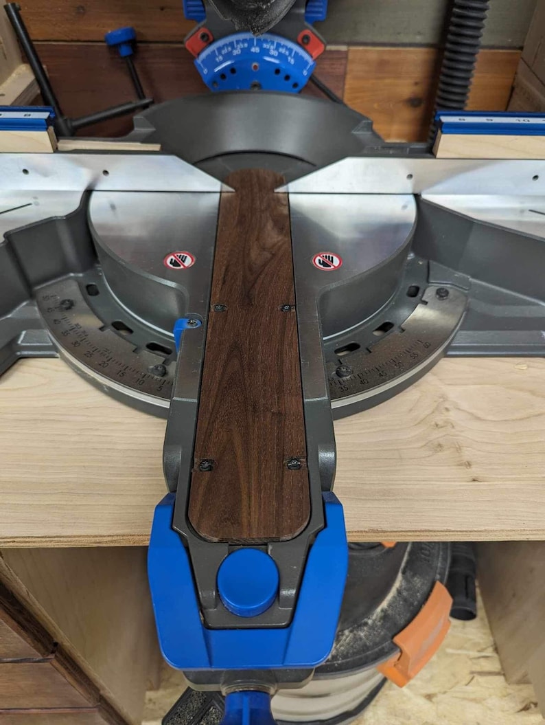 May include: A close-up of a mitre saw with a piece of wood being cut. The saw is blue and black, and the wood is a dark brown colour. The saw has a safety guard and a blade that is visible. The wood is being held in place by a clamp.