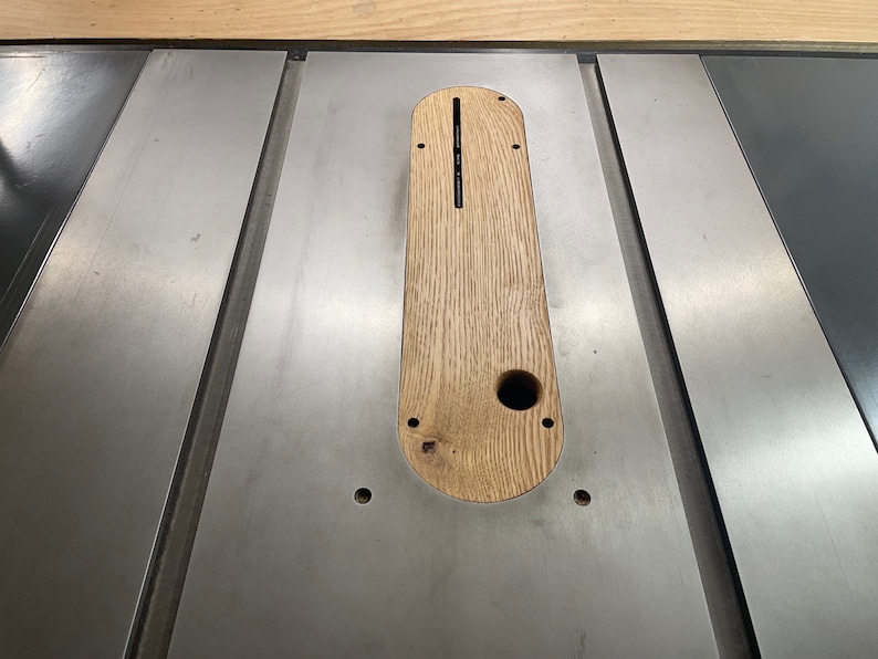 Op de afbeelding: Een close-up van een tafelzaag met een houten inzet in het tafelblad. De inzet heeft een gleuf voor een zaagblad en een gat voor een stofafzuiging.