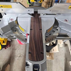May include: A close-up of a miter saw with a piece of dark wood being cut. The saw is silver and has a black handle. The wood is dark brown and has a grain pattern.