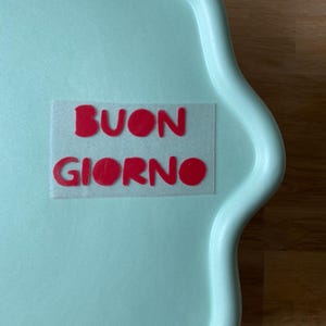Könnte beinhalten: Ein hellblaues Tablett mit einem weißen Aufkleber, auf dem "BUON GIORNO" in roten Buchstaben steht.
