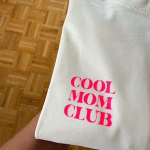 Könnte beinhalten: Off-White-T-Shirt mit dem Text "COOL MOM CLUB" in leuchtendem Pink auf dem Ärmel gedruckt. Das Shirt ist gefaltet und zeigt den Text in einer fetten, serifenlosen Schriftart. Der Stoff scheint ein weiches Strickmaterial zu sein.