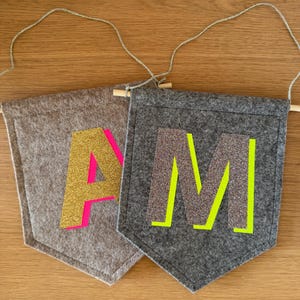 Könnte beinhalten: Zwei Filz-Wimpelbanner mit den Buchstaben "A" und "M". Das "A" ist goldfarben mit pinkfarbener Umrandung, das "M" silbern mit neongrüner Umrandung. Jedes Banner wird an einer Holzstange und Schnur aufgehängt.