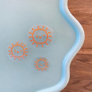 Könnte beinhalten: Drei transparente Aufkleber aus Kunststoff mit orangefarbenen Sonnenmotiven. Jede Sonne hat ein lächelndes Gesicht.