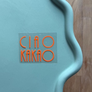 May include: A light teal, scalloped-edge tray with the words "CIAO KAKAO" in bright orange lettering. The tray is made of a solid material and sits on a wooden surface.