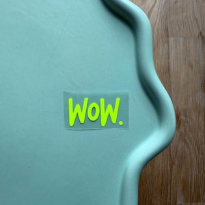 Könnte beinhalten: Ein hellgrünes Tablett mit gewelltem Rand und einem neongrünen Aufkleber mit der Aufschrift "Wow". Der Aufkleber ist mittig auf dem Tablett angebracht. Das Tablett steht auf einer Holzoberfläche.