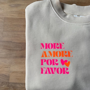 Könnte beinhalten: Beiges Sweatshirt mit dem Schriftzug "MORE AMORE POR FAVOR" in Pink und Orange, mit Herzsymbolen. Das Sweatshirt ist aus weichem Material und liegt gefaltet auf einer Holzoberfläche.