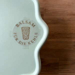 Könnte beinhalten: Ein hellgrünes Tablett mit gewelltem Rand und einem runden Aufkleber. Der Aufkleber zeigt die Worte "BALSAM FÜR DIE KEHLE" um eine Glasgrafik. Das Tablett liegt auf einer Holzoberfläche.