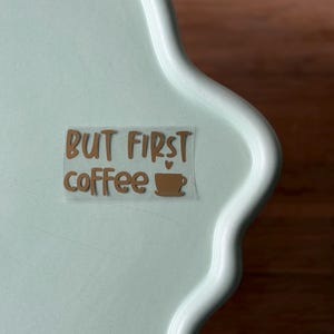 Könnte beinhalten: Ein hellgrünes Tablett mit gewelltem Rand und dem Schriftzug "BUT FIRST coffee" in braunen Buchstaben. Eine kleine braune Kaffeetassen-Grafik befindet sich neben dem Wort "coffee". Das Tablett steht auf einer Holzoberfläche.