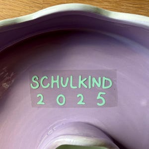 May include: A lavender ceramic bowl with a scalloped rim and a light green edge. The text "SCHULKIND 2025" is printed in light green on a clear sticker in the center of the bowl.