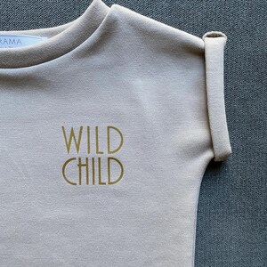 May include: A light beige short-sleeved sweatshirt with the words "WILD CHILD" printed in gold. The sleeve is cuffed. The sweatshirt is made of a soft material.