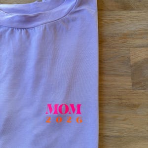 Könnte beinhalten: Ein fliederfarbenes T-Shirt mit dem Wort "MOM" in leuchtendem Pink und den Zahlen "2026" in Orange. Das T-Shirt ist gefaltet, was auf einen neuen Zustand hindeutet. Der Text befindet sich auf der Vorderseite.