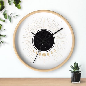 May include: A round wall clock with a light wood frame. The clock face features a black circle with a starry night design, surrounded by gold sunburst rays and moon phases. Black clock hands indicate the time.