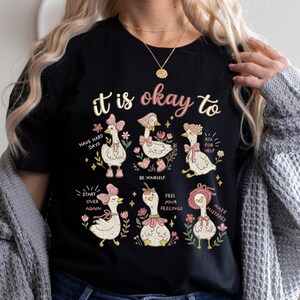以下が含まれることがあります： 「it is okay to」のフレーズがプリントされた黒いTシャツ。6羽の漫画のガチョウのグラフィックが描かれており、それぞれ異なる帽子やリボンを身につけ、花に囲まれています。シャツには「Have hard days」や「Make mistakes」などのフレーズも含まれています。