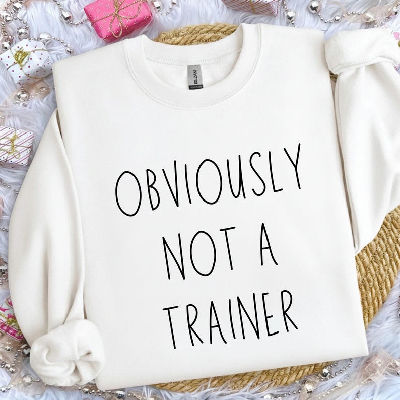 May include: A cream-coloured sweatshirt featuring the black text "OBVIOUSLY NOT A TRAINER". The garment is displayed on a woven surface, surrounded by decorative elements, including wrapped gifts and silver beads. A casual, comfortable item of clothing.