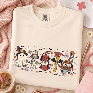 Peut inclure: Un sweat-shirt à col rond couleur crème avec une impression graphique de personnages de dessin animé se tenant la main, incluant un fantôme avec un casque de pompier, une borne d'incendie momie, un extincteur, une hache et un camion de pompiers.