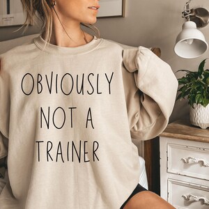 May include: A beige sweatshirt featuring the black text "OBVIOUSLY NOT A TRAINER". The sweatshirt has a crew neck and long sleeves. The image is a close-up of the garment, highlighting the text.