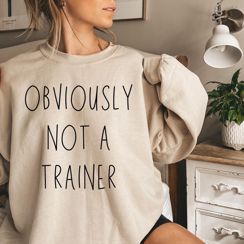 May include: A beige sweatshirt featuring the black text "OBVIOUSLY NOT A TRAINER". The sweatshirt has a crew neck and long sleeves. The image is a close-up of the garment, highlighting the text.