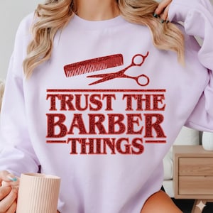 Puede incluir: Sudadera lila claro con un diseño de purpurina roja que presenta un peine y unas tijeras, con el texto "TRUST THE BARBER THINGS" en letras de purpurina roja. Un estilo casual.