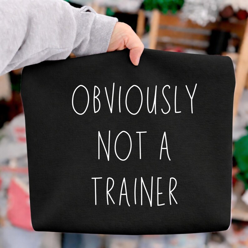May include: A black tote bag with the white text "OBVIOUSLY NOT A TRAINER". The bag is held by a person wearing a grey sweatshirt. The background is blurred, with hints of green and brown.