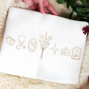 May include: White pillowcase with gold-colored line art of baby footprints, a stethoscope, a swaddled baby, flowers, a heartbeat line, an ID badge, and a clipboard. The pillowcase is held by a hand.