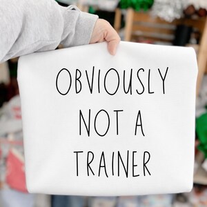 May include: A white rectangular item with the text "OBVIOUSLY NOT A TRAINER" in black, held by a person. The text is in a simple, sans-serif font. The background is blurred, with hints of other objects.