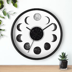 May include: A round wall clock with a black frame and a white face. The clock face features a black and white illustration of the phases of the moon. The center of the clock shows a starry night design.