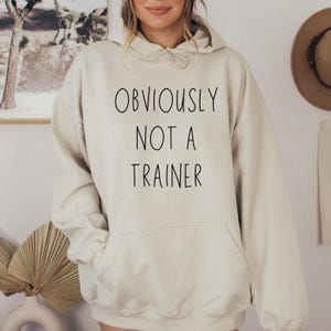 May include: A beige hoodie with the text "OBVIOUSLY NOT A TRAINER" in black. The hoodie features a drawstring hood and a front pocket. The background includes a framed picture and a hat.