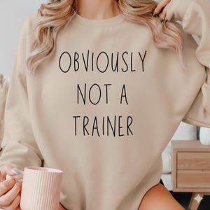 May include: A beige crew neck sweatshirt with the black text "OBVIOUSLY NOT A TRAINER". The sweatshirt is a neutral colour. A pink mug is held in the person's hand.