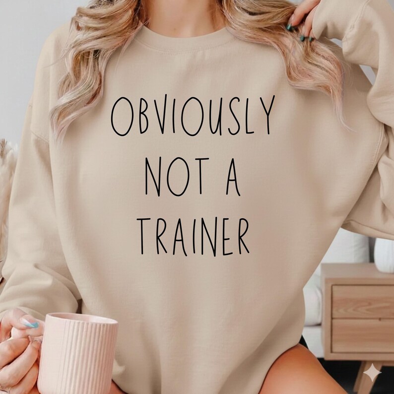 May include: A beige crew neck sweatshirt with the black text "OBVIOUSLY NOT A TRAINER". The sweatshirt is a neutral colour. A pink mug is held in the person's hand.