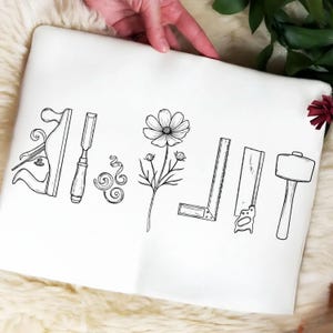 May include: A white fabric item featuring a black line art illustration of woodworking tools including a hand plane, a chisel, wood shavings, a square ruler, a hand saw, and a mallet, centered around a single detailed cosmos flower.