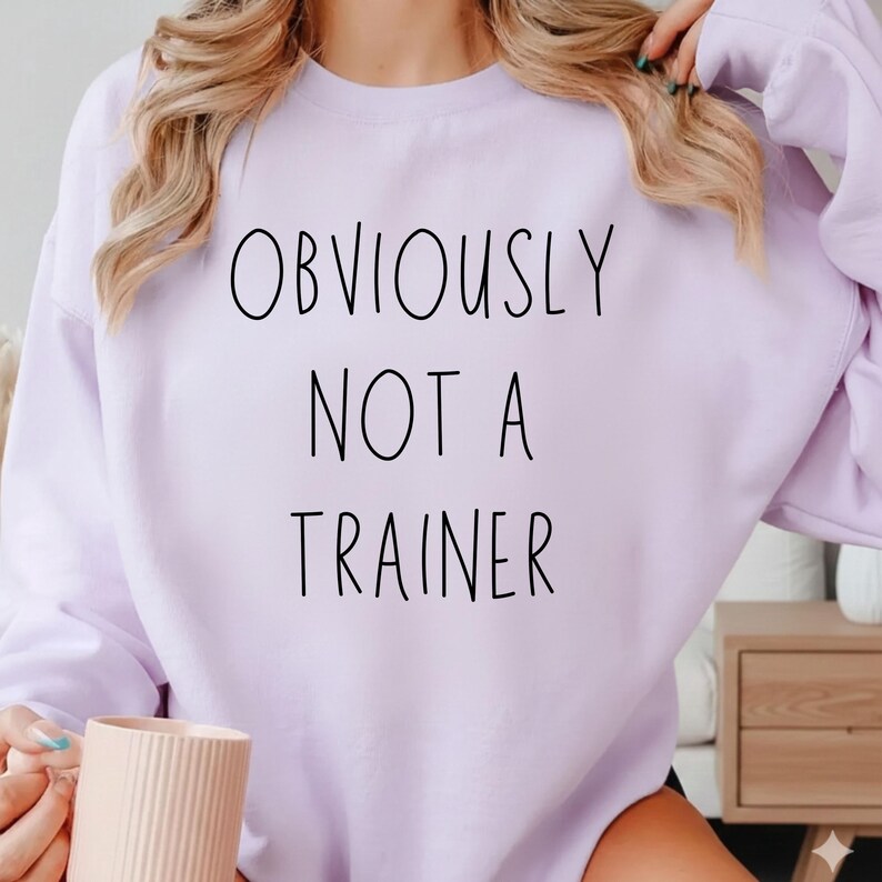 May include: A lilac sweatshirt featuring the black text "OBVIOUSLY NOT A TRAINER". The crew neck sweatshirt is a plain colour. The image shows the upper part of the garment.