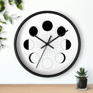 May include: A black and white wall clock featuring a moon phase design. The clock face displays the different phases of the moon in black and white, with black clock hands and a black frame. The clock is round and appears to be hanging on a white wall.