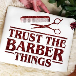 Puede incluir: Cojín cuadrado blanco con texto rojo brillante que dice "TRUST THE BARBER THINGS". Encima del texto hay tijeras y un peine rojos brillantes. El cojín está sostenido por una persona.