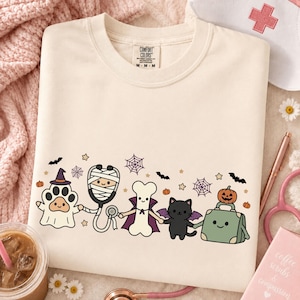Puede incluir: Una camiseta de cuello redondo color crema con un estampado gráfico de cinco lindos personajes de Halloween: un fantasma con sombrero de bruja, un estetoscopio momia, un hueso con capa, un gato negro con alas y un maletín médico verde con una calabaza.