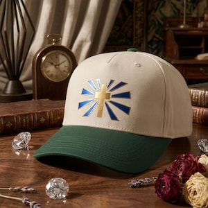 Könnte beinhalten: Eine beige Baseballkappe mit dunkelgrünem Schirm. Auf der Vorderseite befindet sich eine goldene Kreuzgrafik, die von strahlenden blauen Sonnenstrahlen umgeben ist. Die Kappe hat eine strukturierte Krone und einen kleinen grünen Knopf oben.