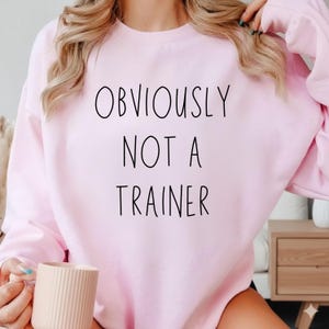 May include: A pale pink sweatshirt featuring the black text "OBVIOUSLY NOT A TRAINER". The long-sleeved top has a crew neck. The image shows the sweatshirt from the chest up.