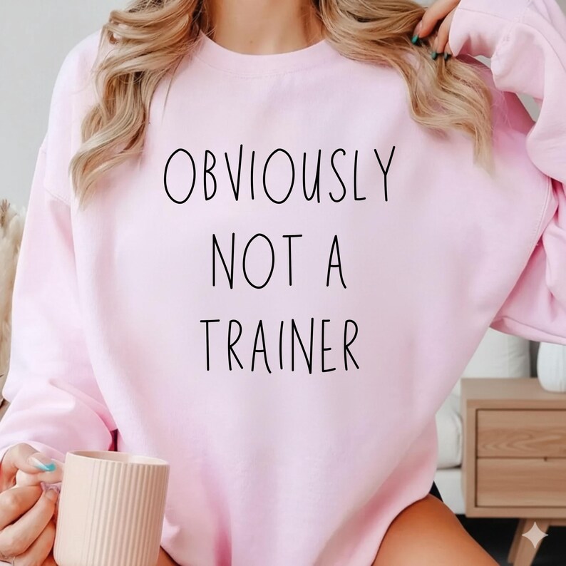 May include: A pale pink sweatshirt featuring the black text "OBVIOUSLY NOT A TRAINER". The long-sleeved top has a crew neck. The image shows the sweatshirt from the chest up.