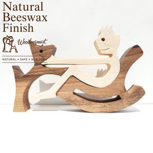 Può includere: Un giocattolo a dondolo in legno con una figura di colore chiaro seduta su una base a dondolo scura, che interagisce con una figura di cane scura. Il testo recita: Natural Beeswax Finish, Woodensmart, NATURAL • SAFE • NON TOXIC.