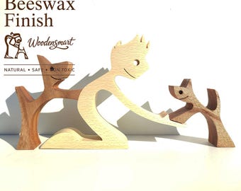 Op de afbeelding: Een lichtgekleurd houten beeldje van een knielende persoon, geflankeerd door twee kleinere donkergekleurde houten hondjes. De tekst "Natural Beeswax Finish", "Woodensmart" en "NATURAL • SAFE • NON TOXIC" staat in de linkerbovenhoek.