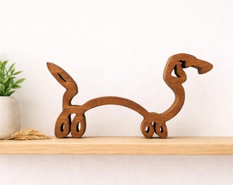 Op de afbeelding: Houten teckel hond sculptuur met een minimalistisch ontwerp. De sculptuur is gemaakt van warmbruin hout en heeft een gebogen lichaam, staart en kop. De hond wordt tentoongesteld op een houten plank.