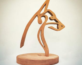 Op de afbeelding: Een houten sculptuur van een hondenhoofd in profiel. De sculptuur is gemaakt van lichtbruin hout en is gemonteerd op een ronde houten basis. De hondenkop is gestileerd met vloeiende lijnen en rondingen, wat een moderne en artistieke esthetiek creëert.