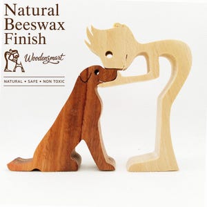 Può includere: Un set di due statuette in legno composto da una figura umana stilizzata di colore chiaro che si china per toccare il naso di un cane in legno scuro. Il testo Natural Beeswax Finish, Woodensmart e NATURAL • SAFE • NON TOXIC è stampato sopra.