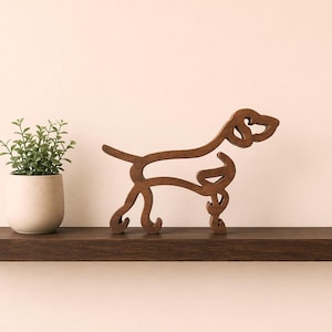 Può includere: Scultura in legno a forma di cane in una calda tonalità marrone. Il design minimalista presenta il contorno di un cane, in piedi su una mensola marrone scuro. Una piccola pianta in vaso con foglie verdi è a sinistra. Arredamento per la casa.
