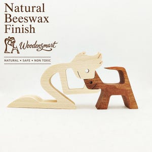 May include: Two wooden figurines featuring a light-coloured person kneeling and a dark-coloured dog. The text reads Natural Beeswax Finish, Woodensmart, and NATURAL • SAFE • NON TOXIC. The figures are crafted from smooth wood with visible grain.