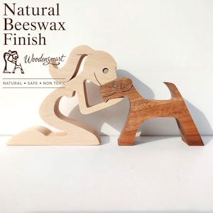 May include: Two stylised wooden figurines featuring a light-coloured figure kneeling and a darker-coloured dog figure standing. The text Natural Beeswax Finish, Woodensmart, and NATURAL • SAFE • NON TOXIC is printed in brown on the upper left side.