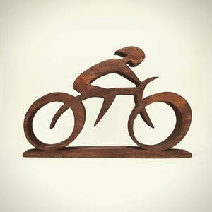 Peut inclure: Sculpture en bois brun fonc&eacute; repr&eacute;sentant un cycliste sur un v&eacute;lo. Le design est minimaliste, avec le cycliste en position de conduite. Les roues du v&eacute;lo sont circulaires, et la sculpture est mont&eacute;e sur une base.