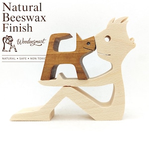 May include: A light-coloured wooden figurine of a person sitting and holding a dark-coloured wooden cat. The text Natural Beeswax Finish, Woodensmart, and NATURAL • SAFE • NON TOXIC is printed in the upper left corner of the image.