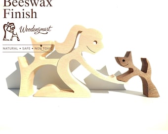 Op de afbeelding: Een set houten figuren in een lichte kleur met een gestileerd persoon die knielt bij twee honden. De tekst Natural Beeswax Finish, Woodensmart en NATURAL • SAFE • NON TOXIC is zichtbaar. De figuren hebben gladde oppervlakken en vormen.