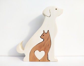 Op de afbeelding: Een houten puzzel van een hond en een kat. De hond is lichtbeige en de kat bruin. Een hartvorm is uit de kat gesneden. De puzzel is gemaakt van gelaagd hout en staat rechtop op een wit oppervlak.